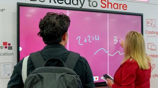 Two ISTE attendees face away from the camera. The woman on the right uses her finger to write a math problem on LG CreateBoard, which is mounted on the wall of the LG ISTE booth, while the man on the left watches.