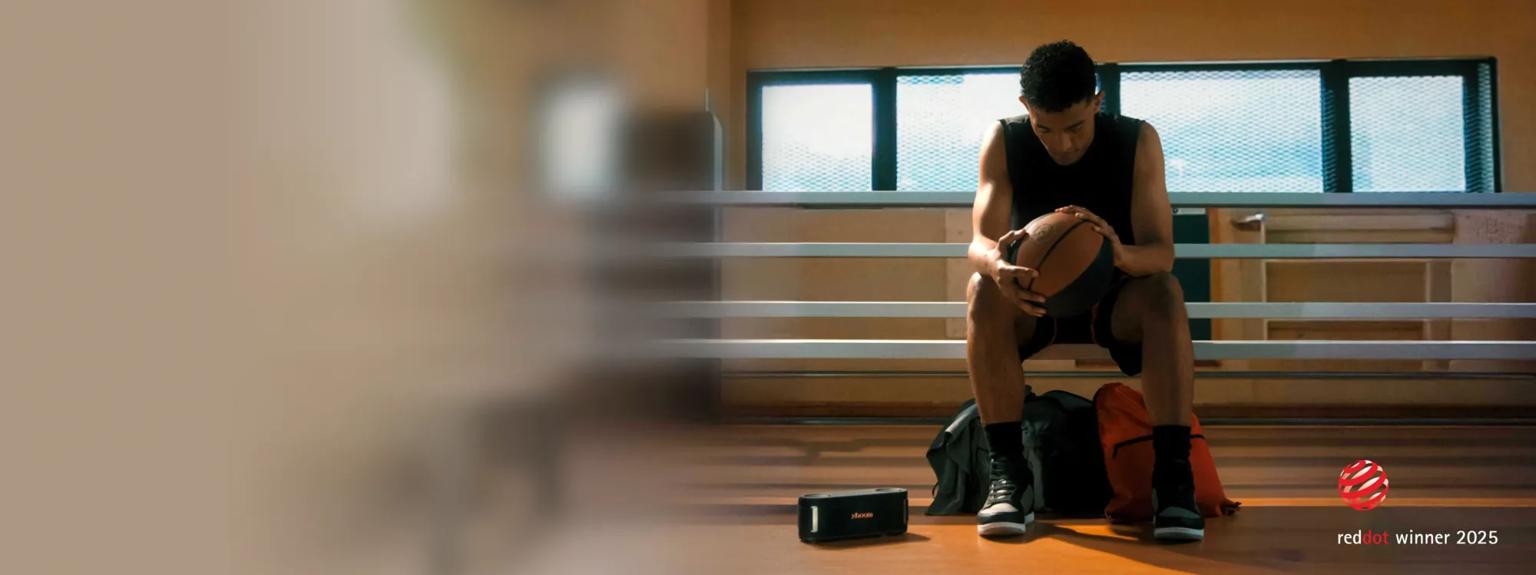 A man rests on a bench while holding a basketball with an xboom speaker nearby. With “reddot winner 2025” badges. 