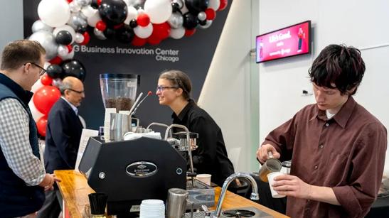 Two baristas make coffee at a stand in an LG Business Innovation Center. One is focused on pouring a latte into a paper cup, while the other barista smiles and chats with several guests attending the BIC event.