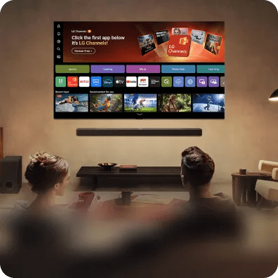 Two people watch a wall-mounted smart TV displaying the webOS interface with various streaming and entertainment apps in a cozy room.
