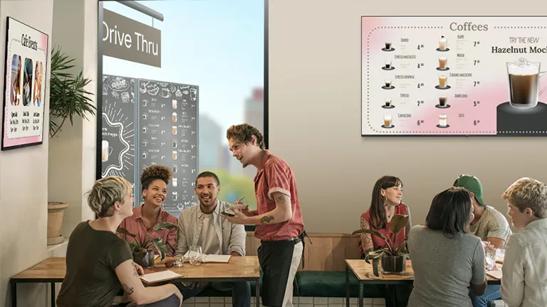 A waiter takes orders from a group at a cafe. LG signage on the walls advertise the coffee menu and upcoming events. 