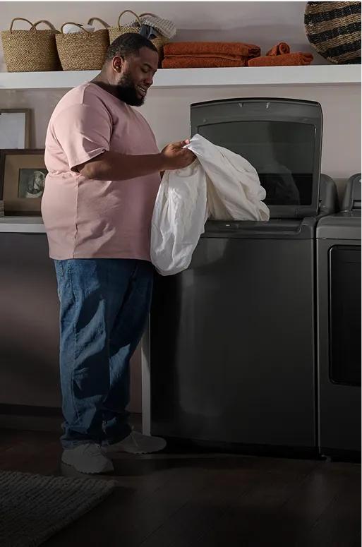 Man removing a white sheet from a dark gray top-load washer.
