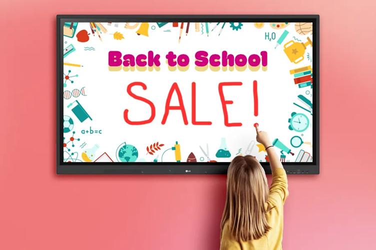 An LG CreateBoard displaying the words Back to School with a colorful border of items like rulers, globes, apples, and other school items. An elementary school student reaches up to write the word Sale! below it using the touch screen, so the message reads Back to School Sale!