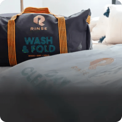 Dry cleaning garment bags and a bag for Rinse's wash & fold service sit on a bed.
