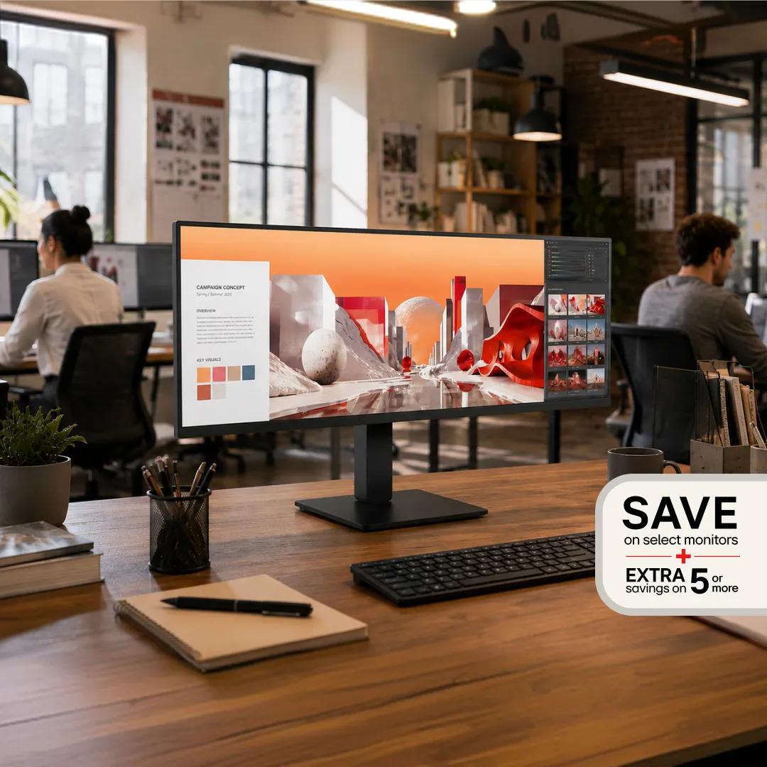 An ultrawide LG monitor on a wooden desk in an open office, displaying a creative campaign concept, with a badge reading 'Save on select monitors, extra savings on 5 or more.'