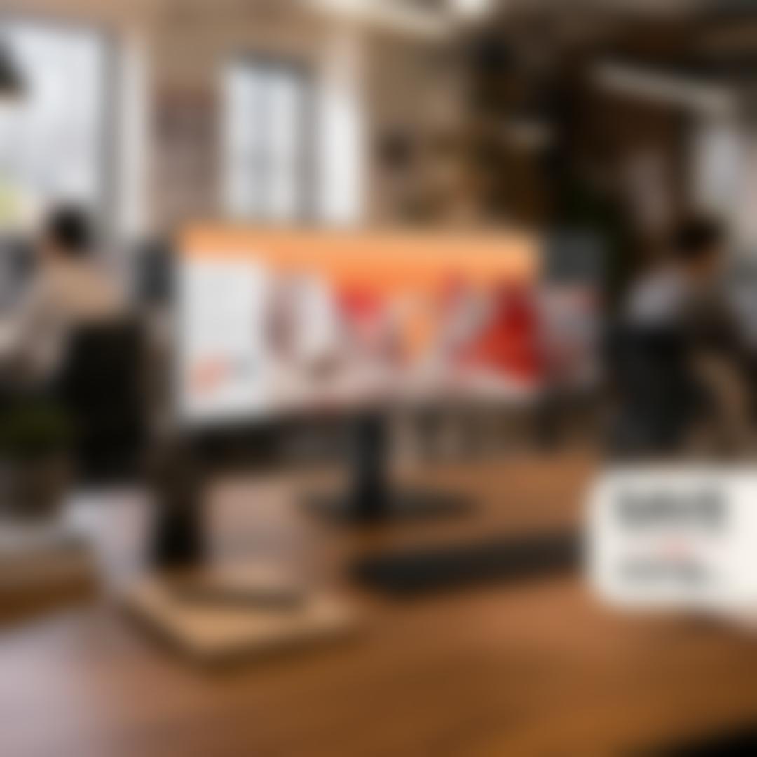 An ultrawide LG monitor on a wooden desk in an open office, displaying a creative campaign concept, with a badge reading 'Save on select monitors, extra savings on 5 or more.'