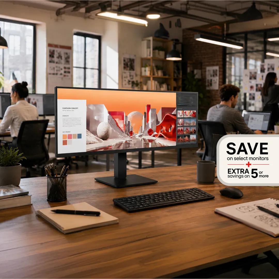 An ultrawide LG monitor on a wooden desk in an open office, displaying a creative campaign concept, with a badge reading 'Save on select monitors, extra savings on 5 or more.'