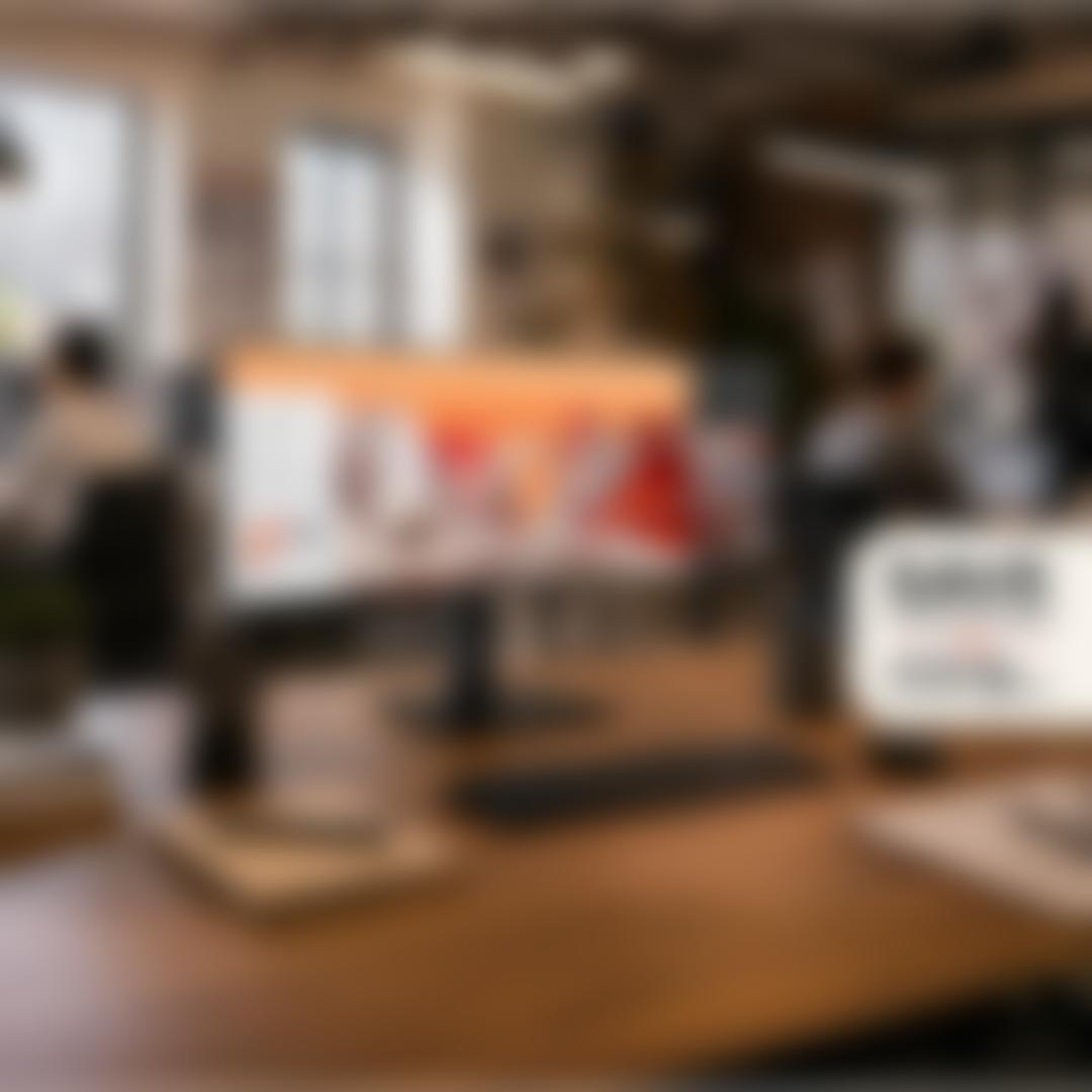 An ultrawide LG monitor on a wooden desk in an open office, displaying a creative campaign concept, with a badge reading 'Save on select monitors, extra savings on 5 or more.'