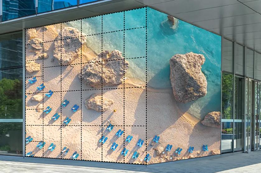 A large digital screen on a building shows an aerial beach scene with rocks and blue chairs.