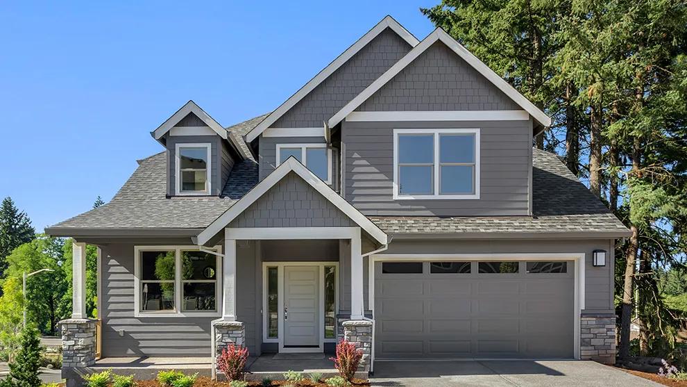 Exterior view of a two-story single-family home with gray siding, stone accents, covered front porch, and attached two-car garage surrounded by trees under a clear blue sky.