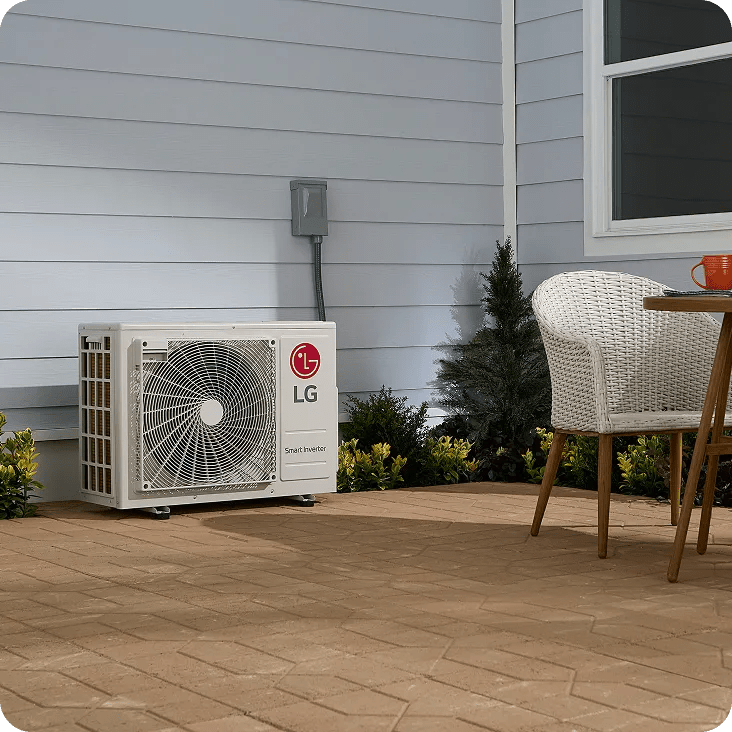 Compact LG outdoor HVAC unit installed on a brick patio next to a white-paneled house, with nearby shrubs, a wicker chair, and a table with an orange cup.