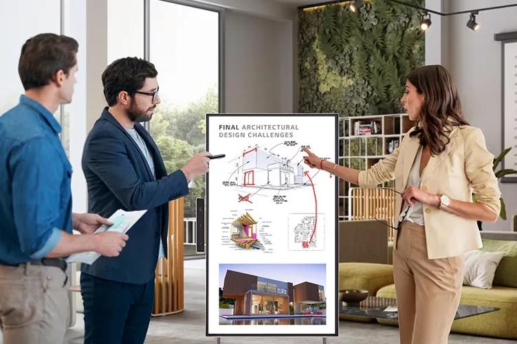 Three professionals stand in a modern living room or office space, looking at a One:Quick Flex. Displayed on the One:Quick are images of architectural drawings and a photo rendering of a modern house. Arrows and other markings in red digital ink show the touch screen has been utilized to annotate the images.
