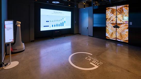 Entry space of the LG LA Business Innovation Center, with the LA BIC logo displayed on the floor via projector. To the left is a large digital display showing sample charts; to the right are the glass doors leading to the BIC, with transparent OLED showing a bright pattern.