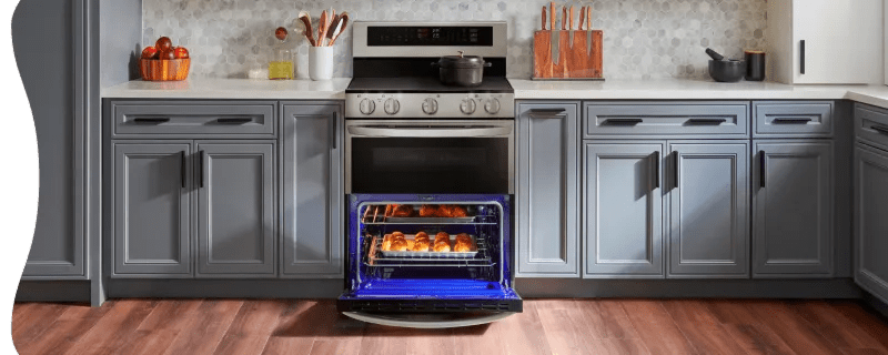 A modern kitchen features a stainless steel LG range with an open oven door revealing a bright blue interior and baking pastries. The setup includes a matching over-the-range microwave, two-tone grey and white cabinetry, and a grey hexagonal tile backsplash.