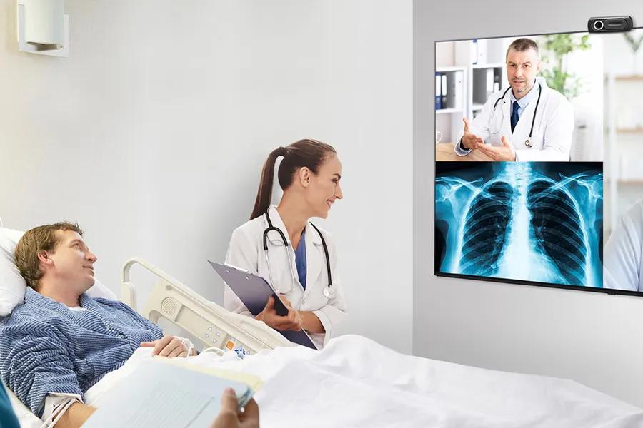 A doctor sits besides a patient reclined in a hospital bed. They're both looking at an LG Pro:Centric Smart Hospital TV, with another doctor on a video call and an x-ray both visible on the display. 
