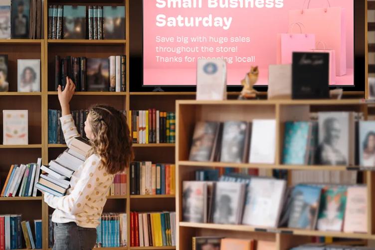A woman holding a stack of books in a bookstore selects another from the shelf. An LG Professional TV on the shelf beside her shows a Small Business Saturday graphic adverting store deals.