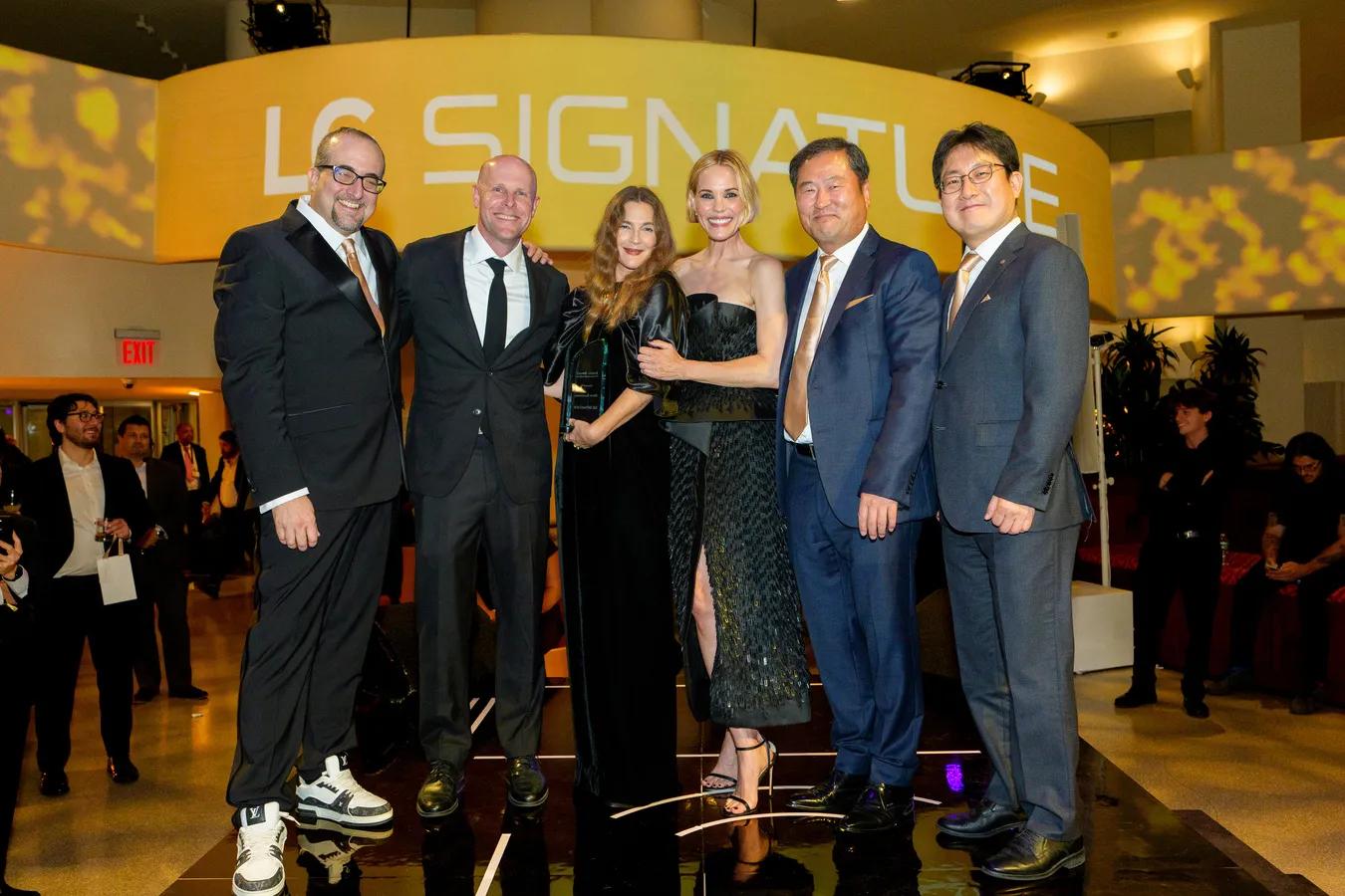 Six people in formal attire pose together on a stage in front of a yellow ‘LG Signature’ sign, with one woman holding a glass award and guests mingling in the background.