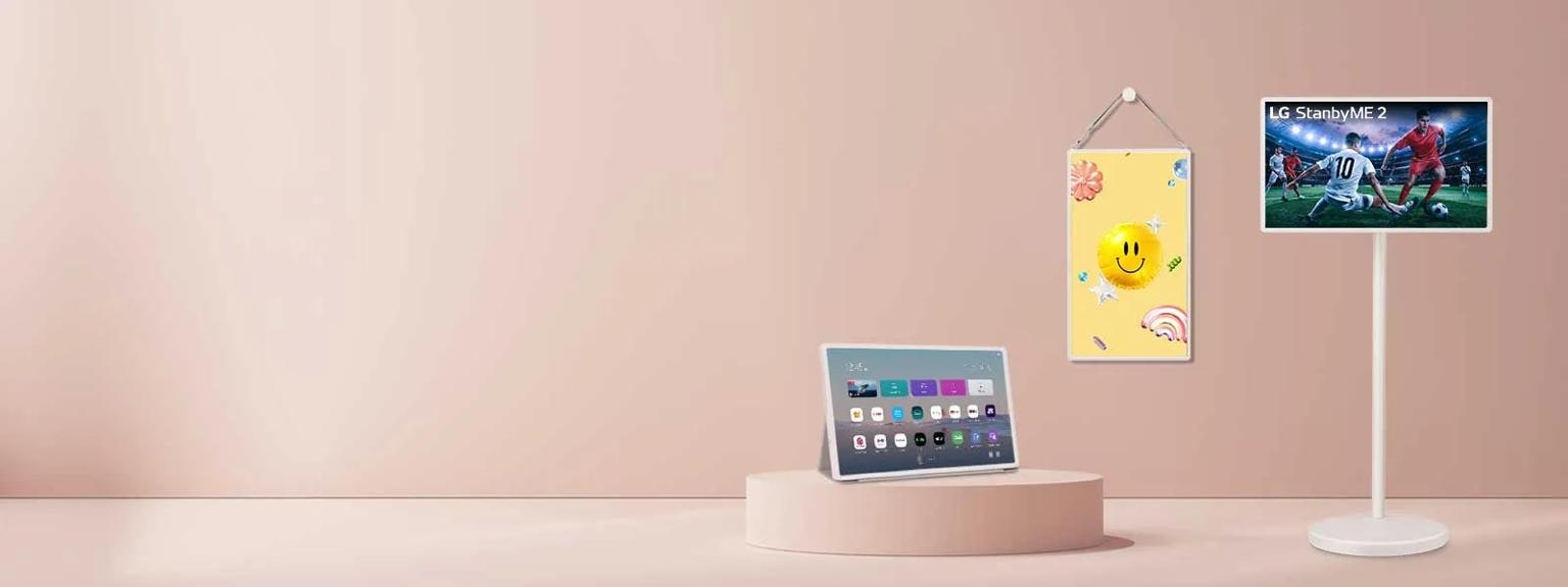 An image featuring three LG StanbyMe 2 smart displays—on a stand, hanging on the wall in portrait orientation, and as a tablet in a folio case set on a pedestal—arranged against a soft pink backdrop. Each device showcases different vibrant content, ranging from live sports to a playful smiley face, emphasizing a versatile and minimalist tech aesthetic.