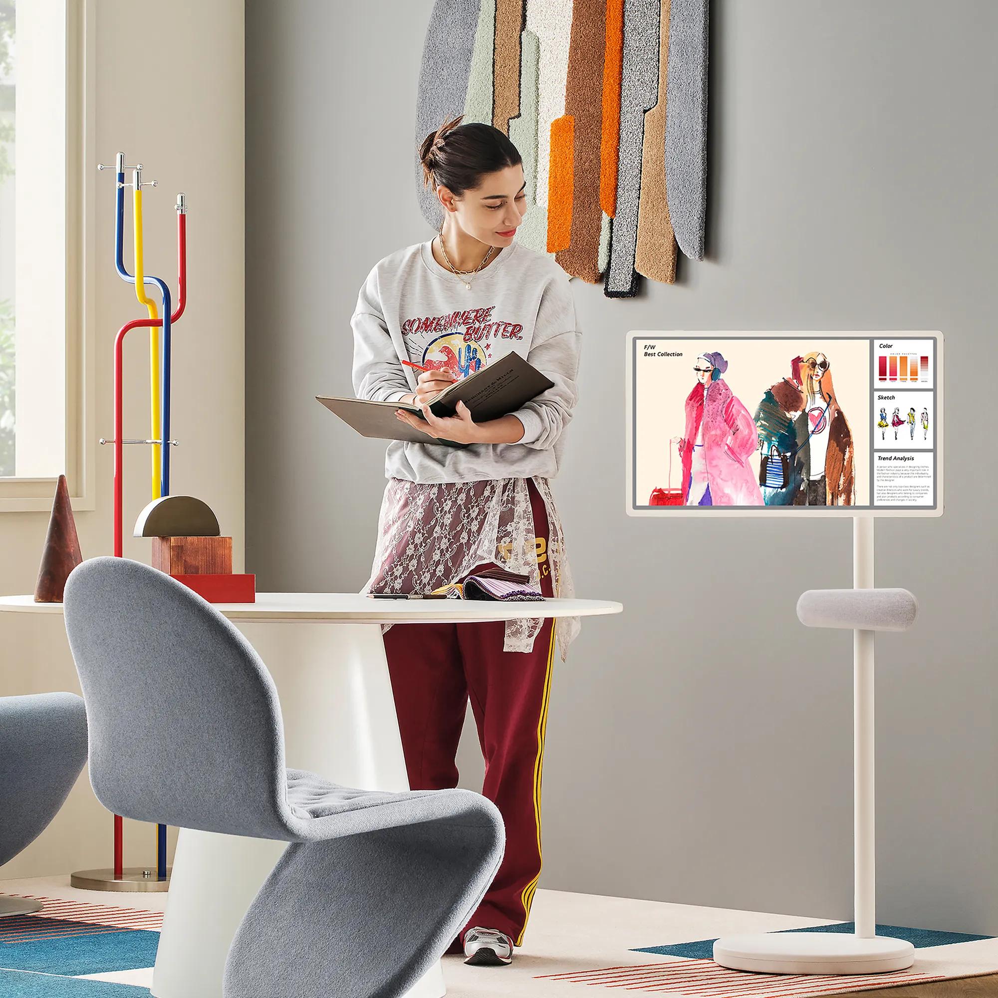 Woman standing in a stylish living space, holding a notebook and viewing fashion sketches displayed on LG StanbyME 2 on a standing display.