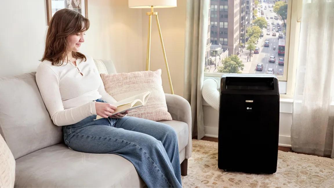 Fall Savings -- LG portable air conditioner in a living room beside a window, with a woman reading on a sofa nearby