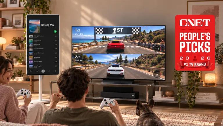 Two people and a dog in a living room playing a split-screen racing game on a large LG TV with Xbox controllers, featuring a vertical smart display to the side and a CNET People's Picks 2026 #1 TV Brand badge.