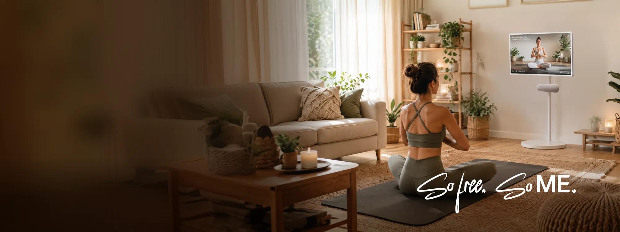 Woman practicing yoga on a mat in a warm living room, facing an LG StanbyME display on a floor stand showing a yoga tutorial, with text reading: So free. So ME.