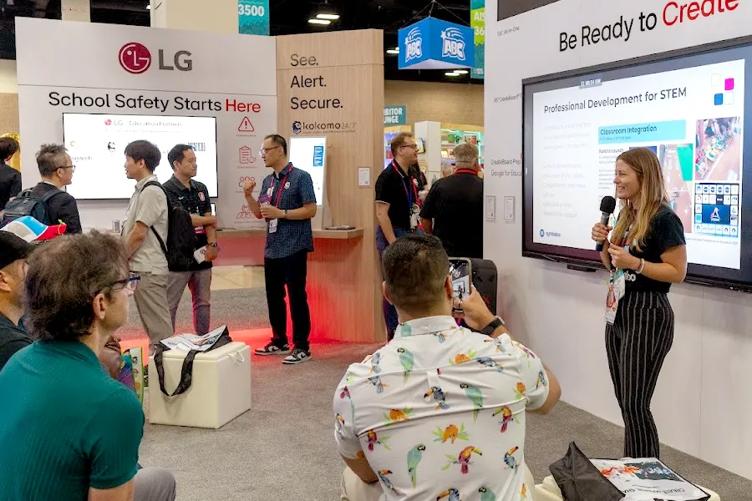 A woman gives a presentation at the LG ISTE booth to a rapt audience. She's holding a microphone in one hand, and behind her is an LG CreateBoard for use in her presentation. In the background, other attendees chat and explore LG solutions at the booth. 