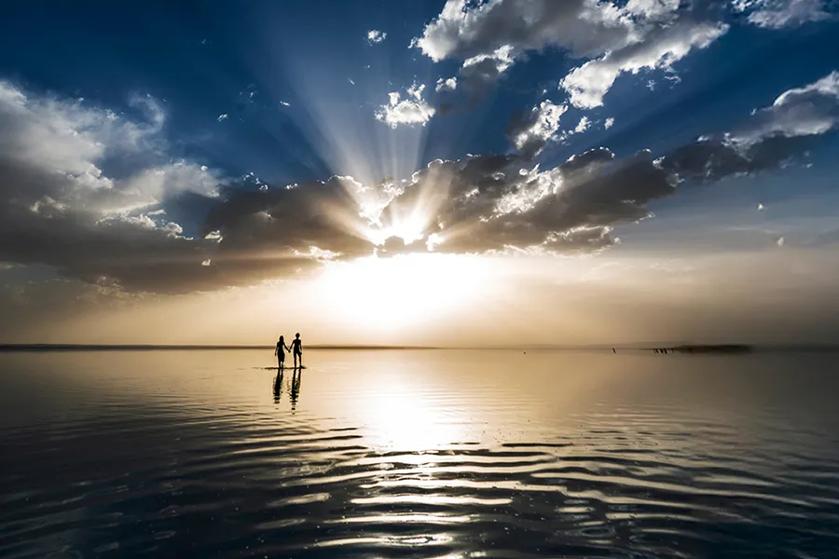 Two people stand on a reflective body of water, holding hands against a dramatic sunset sky with rays of light.