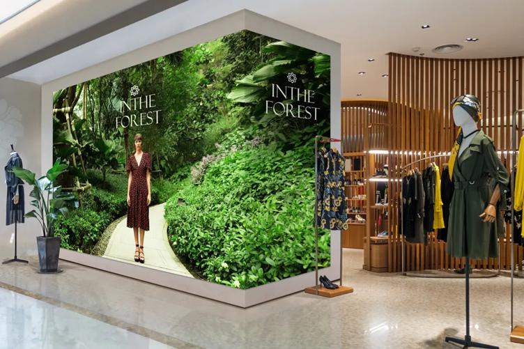 An indoor mall scene with a boutique, upscale clothing store. One wall has bold, vivid indoor Direct View LED signage showing a woman in a sundress on a forest path.