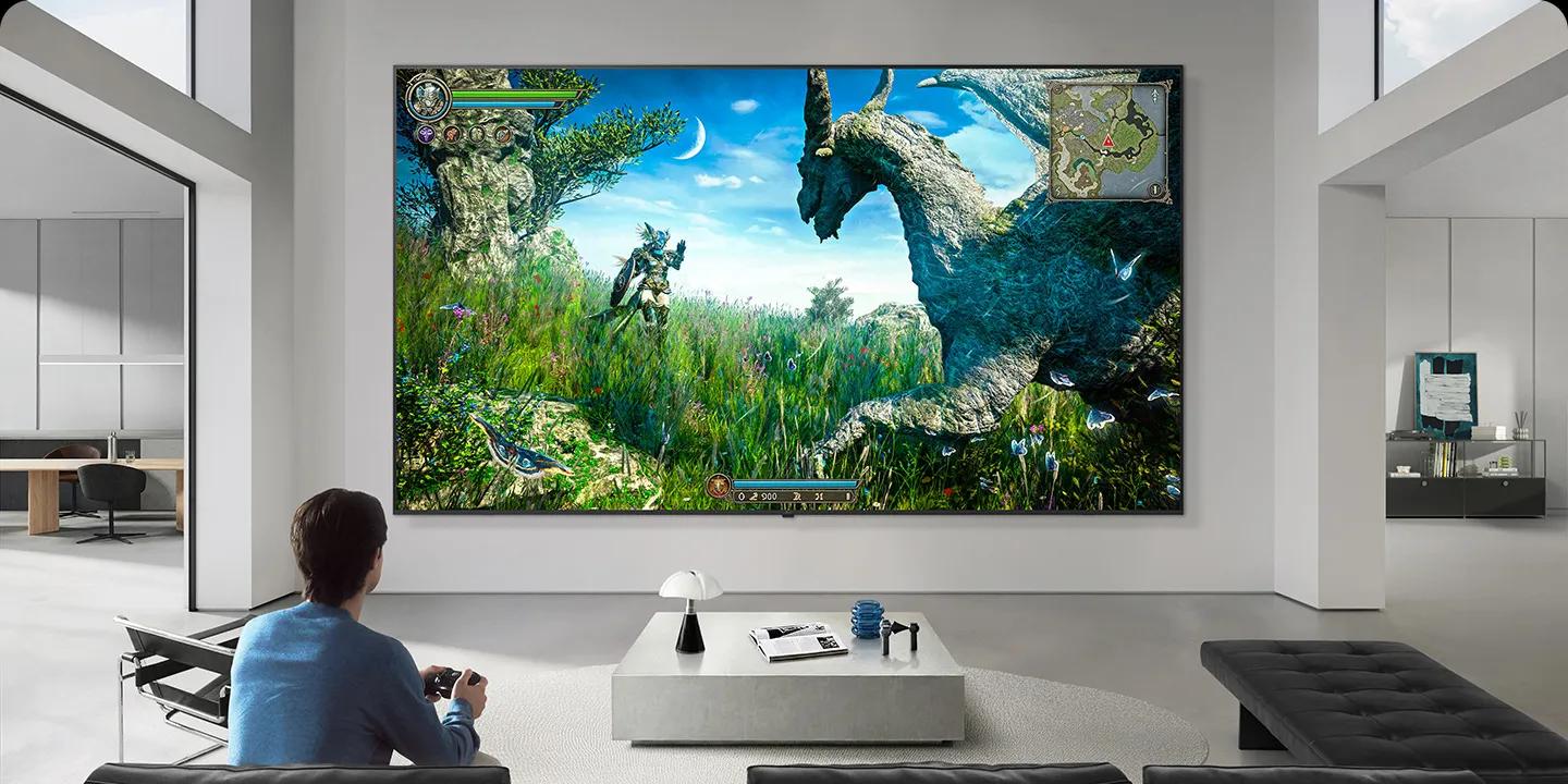 LG TV appears mounted on a wall in a black-and-white-toned living room, showing a fantasy dinosaur game scene on a large display while a person sits on a sofa holding a controller.