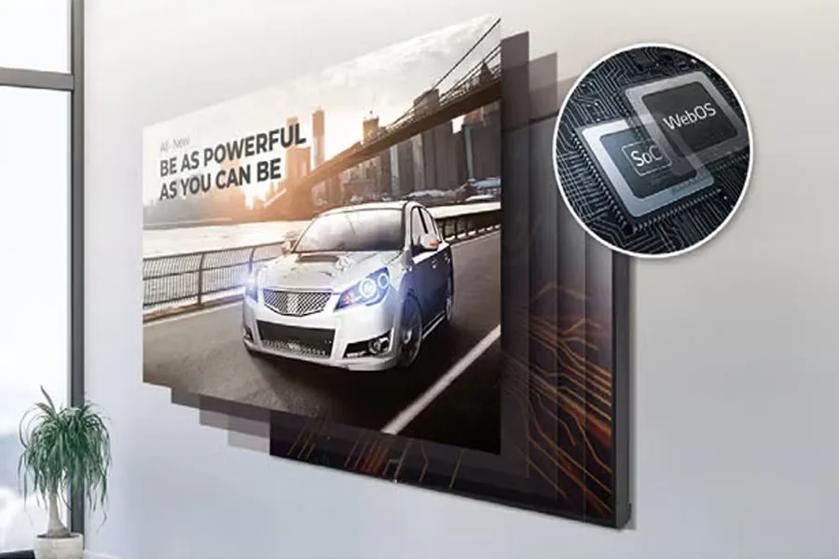 The image displays a high-tech advertisement for an automotive or technology company. At the center, there is a large flat-screen display mounted on a wall showing a silver car on an urban road. The backdrop of the display features a city skyline with a prominent bridge under a sunset glow. The car has glowing headlights, suggesting advanced technology or power. In the top right corner, there is an inset magnified view of microchip-like components labeled "SoC" and "WebOS," indicating digital or software features. The room has minimal decor, with the edge of a plant visible on the bottom left corner, emphasizing a clean, modern aesthetic.