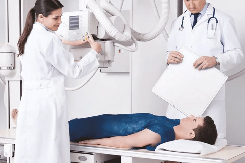 Two calm and smiling doctors stand on either side of a patient, who is lying on an X-ray table. One doctor is moving the x-ray imager over the patient. The other is holding an LG digital x-ray detector.