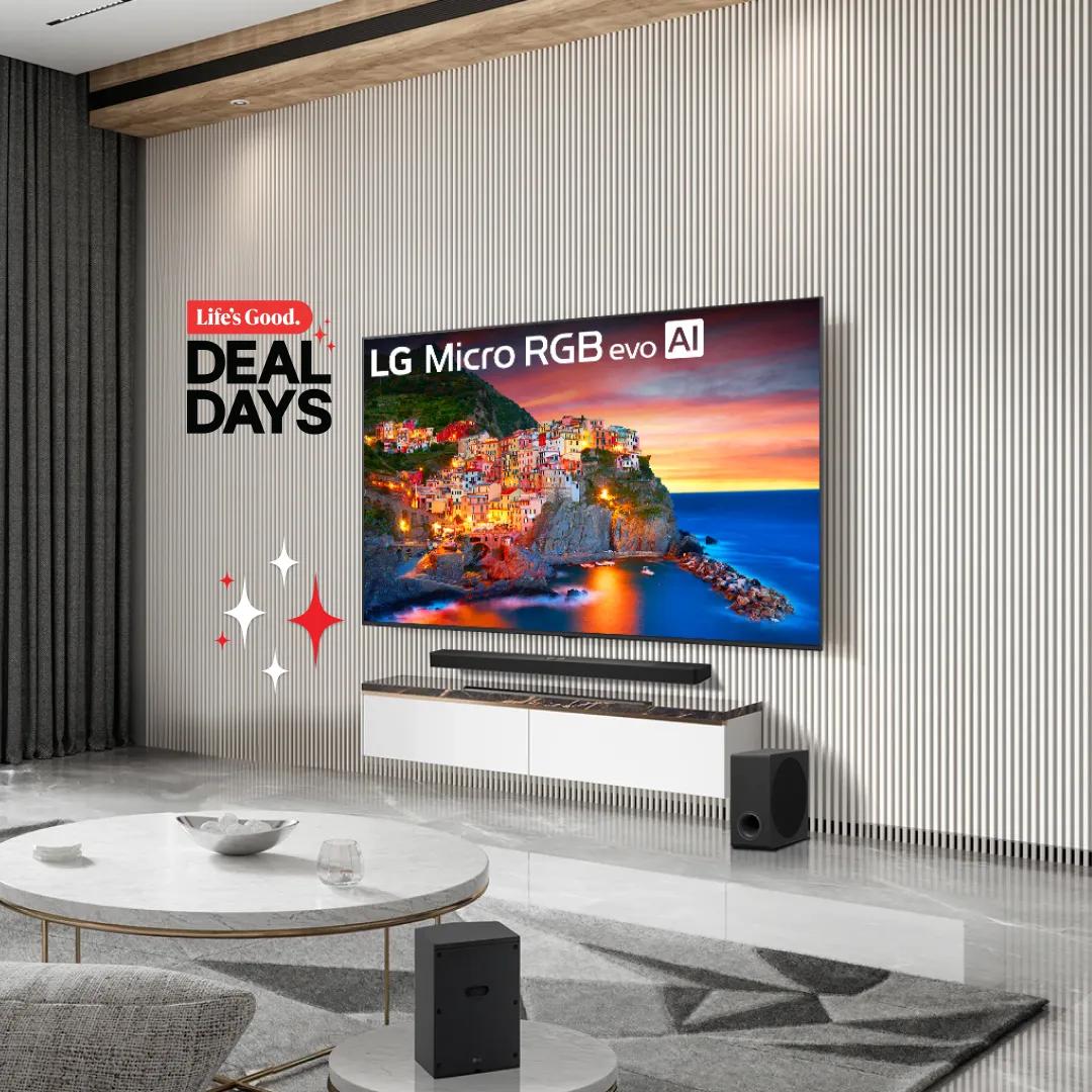 A large LG Micro RGB evo AI TV hangs on a slatted white wall over a floating white media console. The screen shows a colorful cliffside village above the sea at sunset. A soundbar sits under the TV, with a subwoofer beside the console and a rear speaker in the foreground near a marble coffee table. Red and white sparkles surround the “Life’s Good Deal Days” logo on the left.
