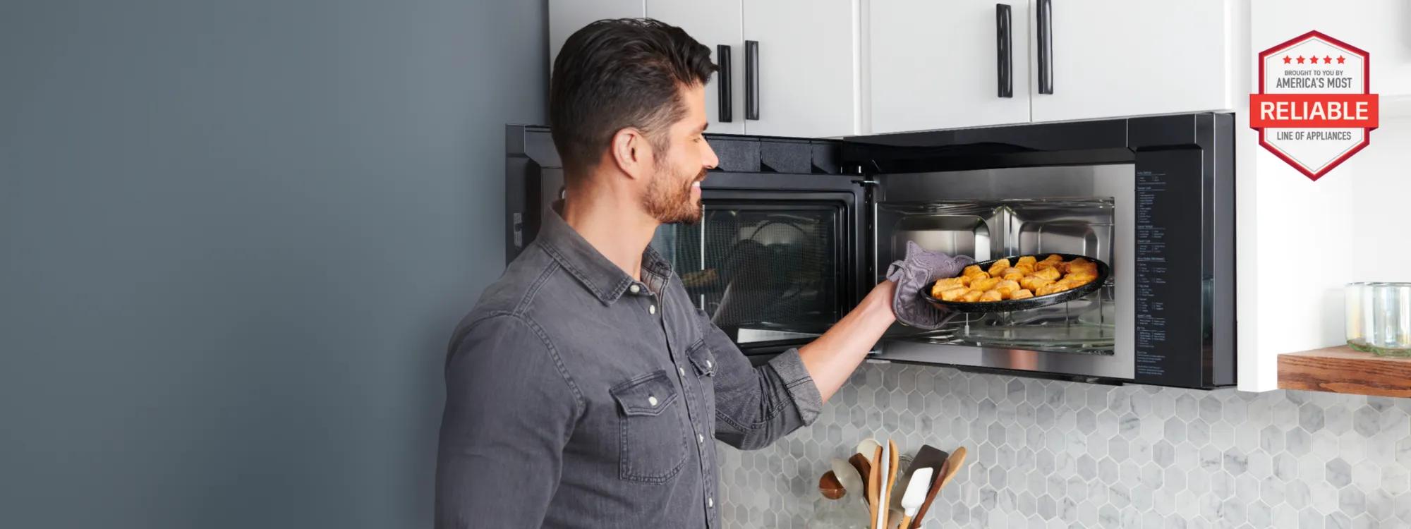 Man slides a plate of food from an LG over-the-range microwave in a bright kitchen, with white cabinets, tile backsplash and black trim above.