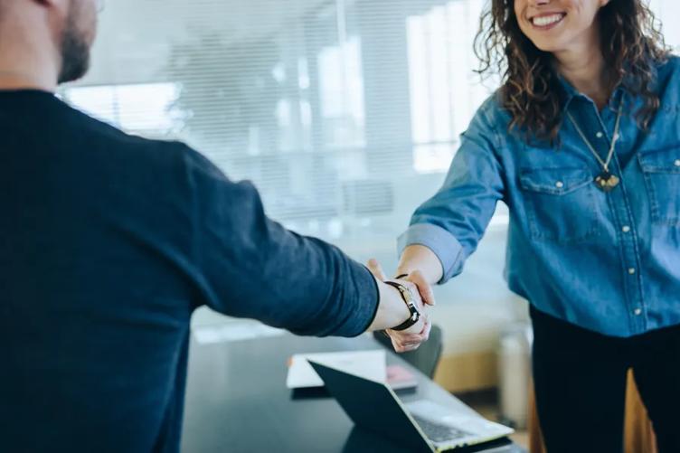 A smiling woman reaches across her desk to shake hands with a man. 