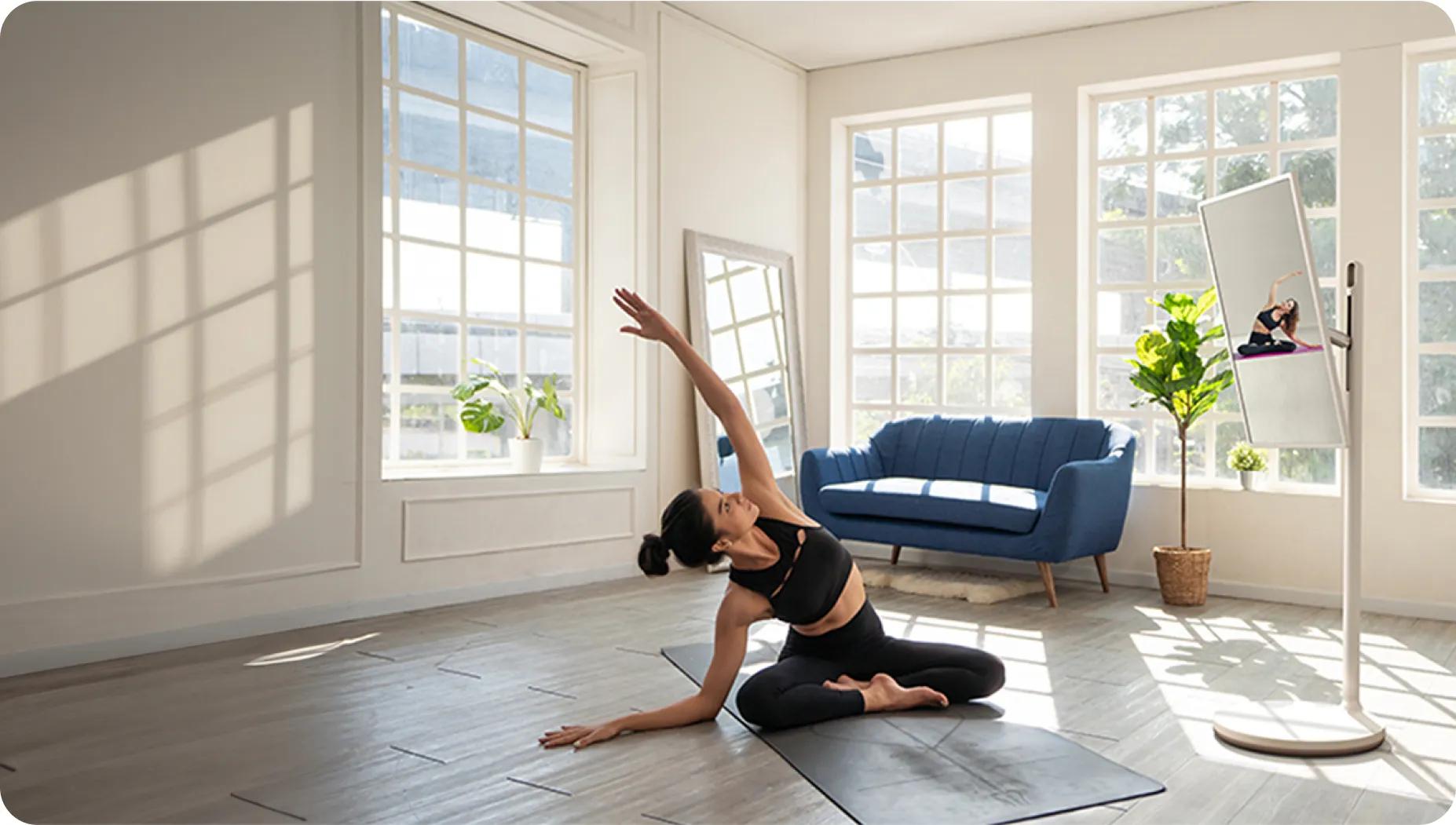 Bright sunlit room with woman stretching on yoga mat watching a workout video on an LG StanbyME on a portable stand.