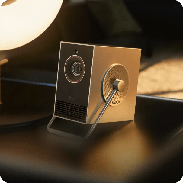 An LG CineBeam Q projector on a table, shown from the side and front with its lens and support stand visible, lit by warm ambient light.