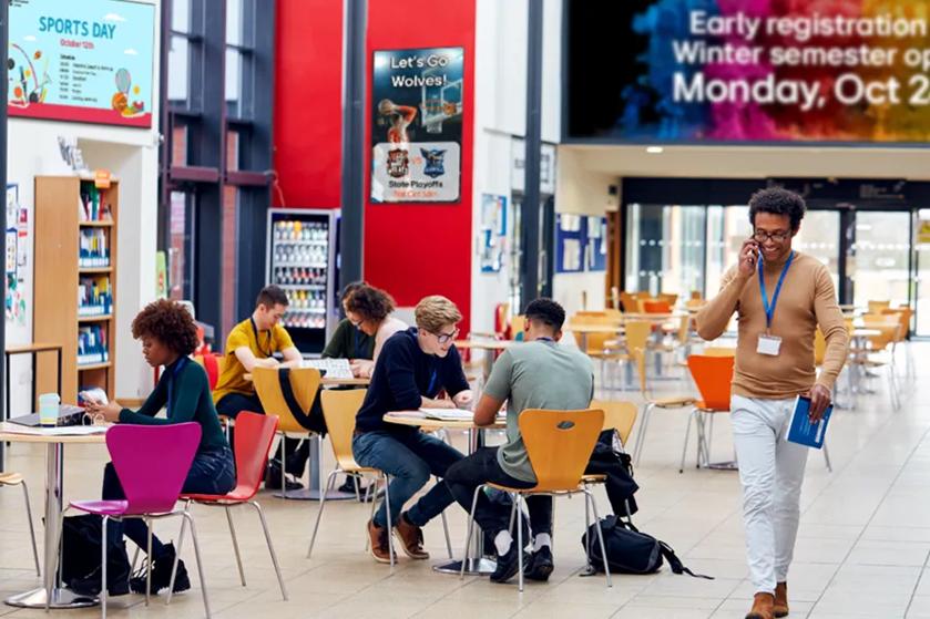 University cafeteria with students eating, studying, and socializing. LG digital signage and DVLED advertise school events and enrollment deadlines.
