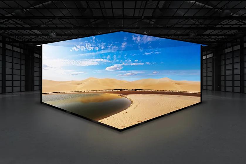 A large, angular screen showing a desert landscape with sand dunes and a small oasis inside a spacious industrial room.