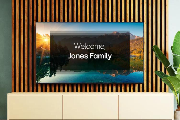 An LG hotel TV is mounted above a modern sideboard with wood paneling behind it. The display shows a nature scene, overlaid with text reading Welcome Jones Family.