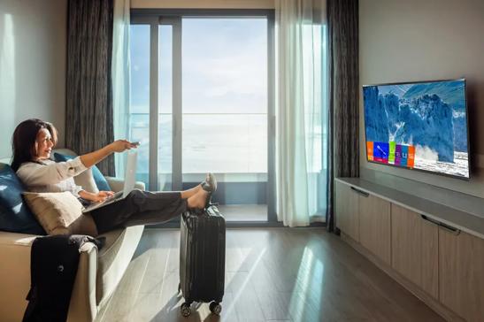 A businesswoman sitting in a cruise cabin, on the couch with her feet propped on her suitcase. She's pointing a remote at the LG Cruise Cabin TV on the wall opposite her. The TV shows a icy glacier scene with the Pro:Centric user menu overlaid on it. Behind her, a large window shows the open sea.