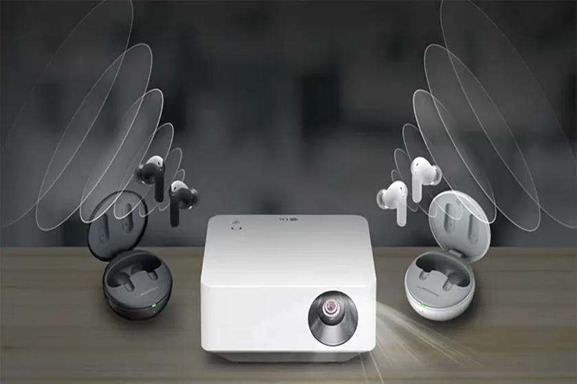 The image depicts a sleek, modern setup of electronic devices on a wooden surface. At the center is a rectangular, white projector with a lens on the right side. To the left, there is an open round case containing a pair of black earbuds, with the case displaying a small green indicator light. To the right, another open round case holds white earbuds, also with a similar green indicator light. Above the earbuds, translucent, oval-shaped sound waves are illustrated, emanating outward to suggest sound in motion. The background is blurred, ensuring focus remains on the electronic devices.