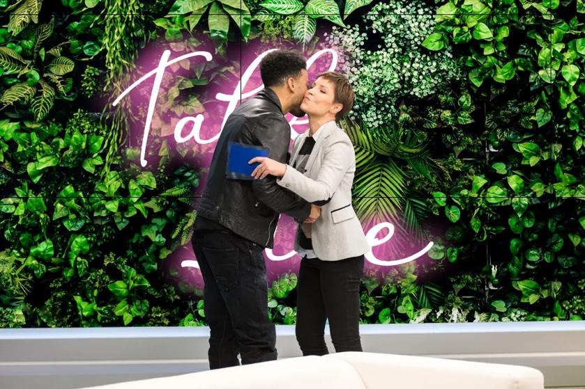 A talk show host embraces her guest. Behind them is a large LG Video Wall with a greenery background and pink script reading Talk Live!
