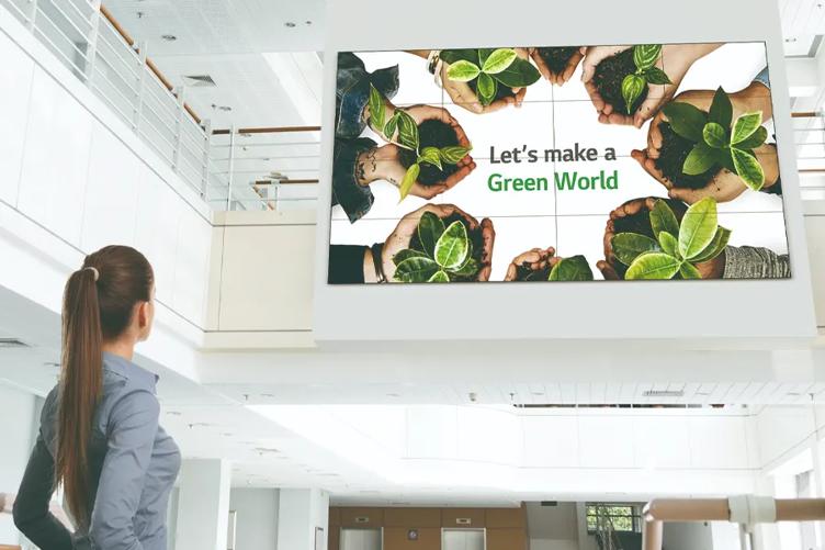 A woman in a large lobby space looks up at the LG video wall mounted in the central area. On it, several pairs of hands hold bright green plants in soil, with the words 'Let's make a green world' in the middle. 