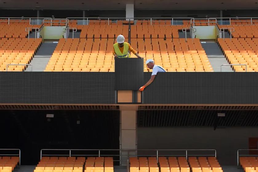 Two construction workers installing a digital screen in a stadium with orange seats.