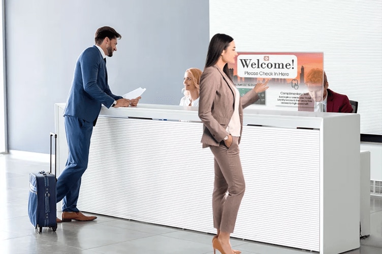 Two business people checking in at a modern hotel lobby. The man in the background is speaking to the receptionist. The woman in the foreground is using a transparent touch OLED display mounted to the desk to check herself in. 