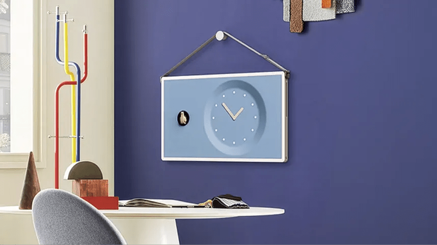 LG StanbyME 2 displayed as a digital picture frame on a purple wall, showing a clock face illustration in a modern workspace.