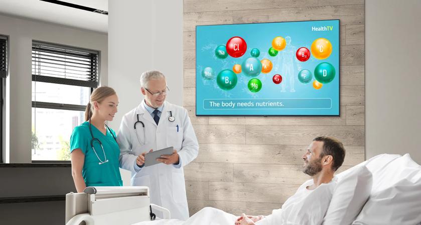 A hospital room with a patient in bed speaking to two medical professionals. A screen on the wall shows nutrient symbols and the text "The body needs nutrients."
