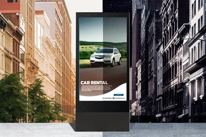 The 75XE3C outdoor display with a graphic for a car rental service stands on a city street. The scene is divided into a day half and a night half. The display is brighter on the day half and dimmer on the night half of the image, demonstrating the 75XE3C's smart brightness control.