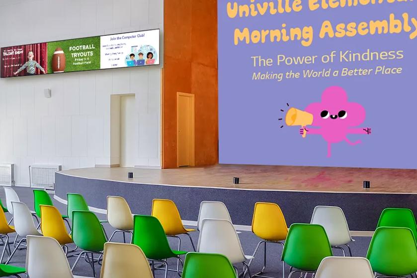 A school auditorium with rows of bright colored chairs before a stage. LG DVLED Display forms a backdrop displaying a message about the morning assembly. On the wall nearby, stretch signage shows upcoming school events.